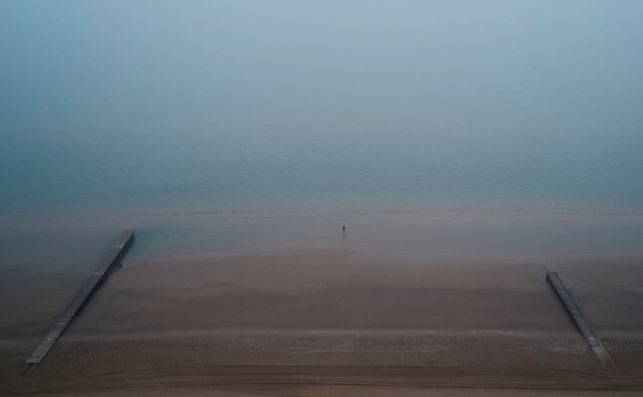 Man standing on a sandy beach, in the middle of two pier, in a foggy day.